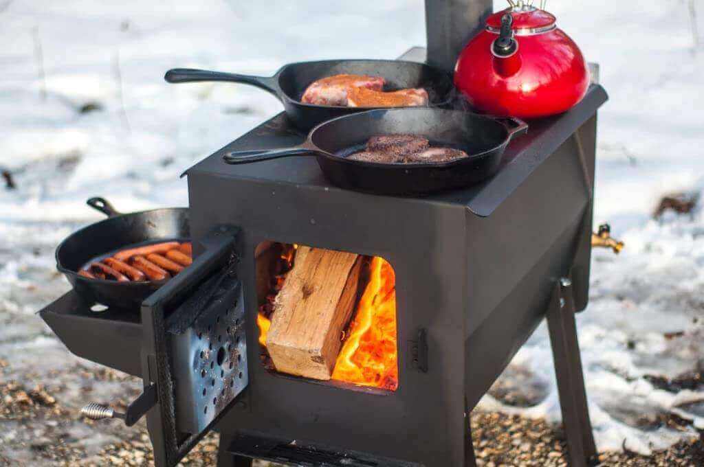 Englander Black Bear Camp Stove from Cabin Depot with logs burning, topped with pans and a teapot, ideal for camping.