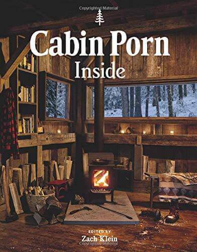 Cozy cabin interior from 'Cabin Porn Inside' book by Cabin Depot, showcasing a rustic wood-burning stove and forest view.