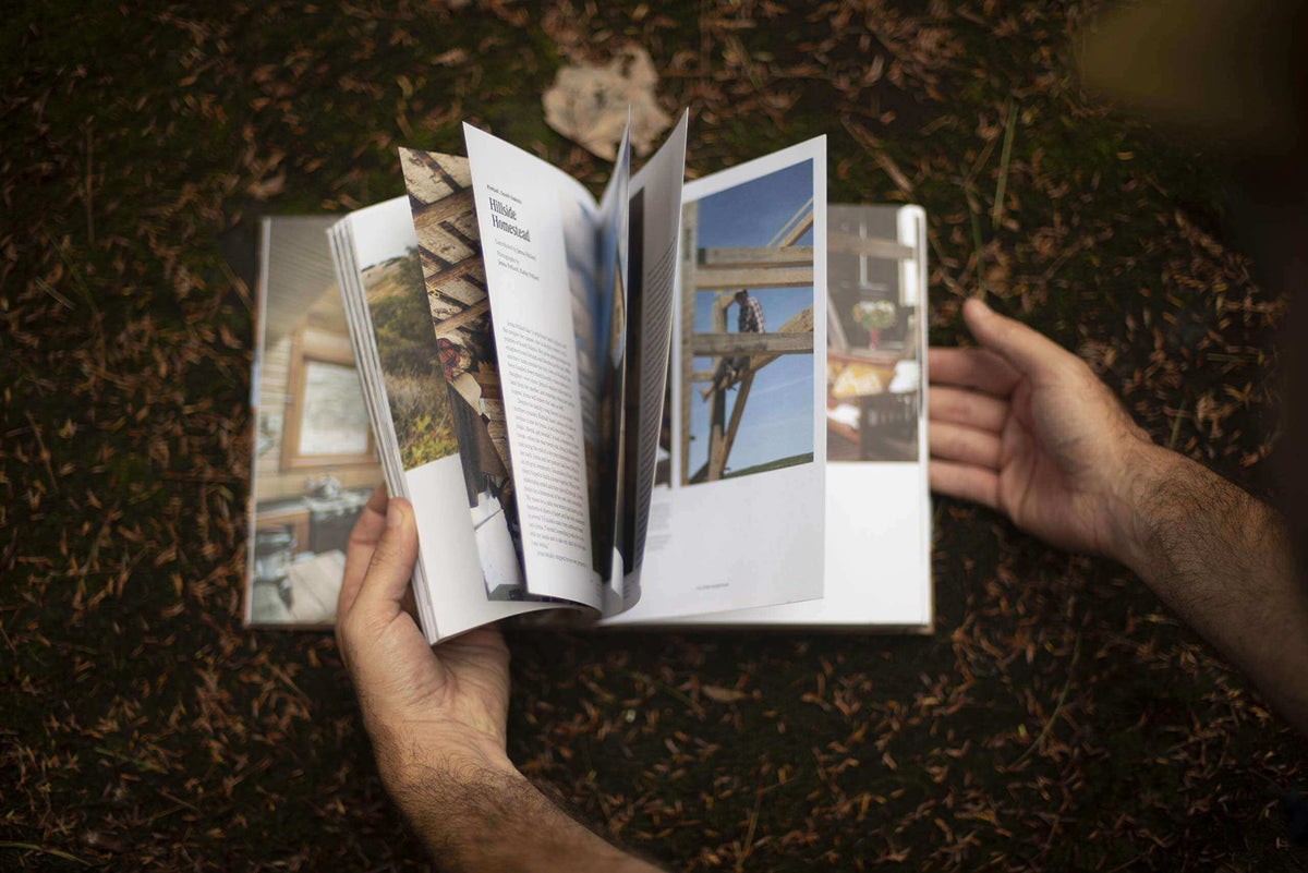 Man leafing through Cabin Porn Inside book in nature, showcasing tranquil cabin images. Available at Cabin Depot for homebuilding inspiration.