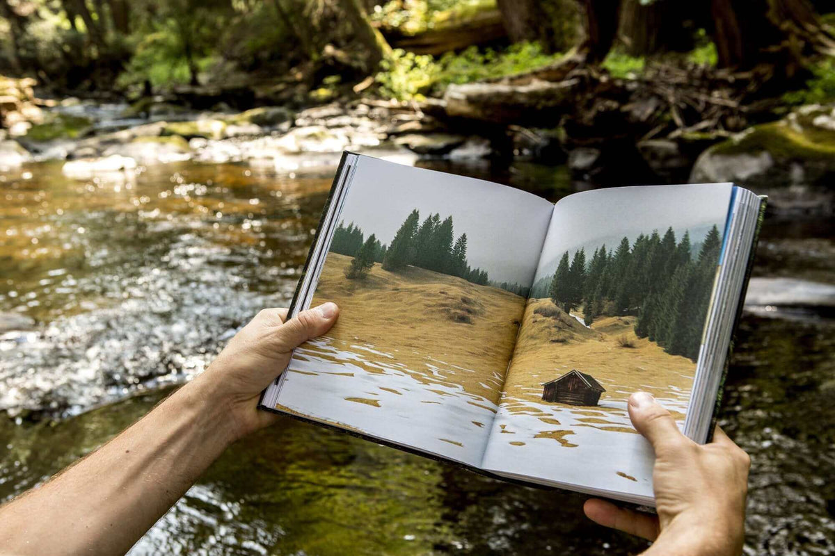 Hands holding 'Cabin Porn' book by a forest stream, featuring serene cabin and nature images. Perfect rural escape inspiration. Cabin Depot.